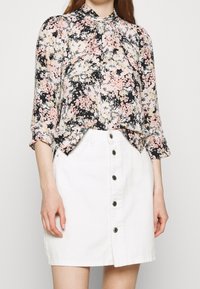 Blusa floreale in tessuto nero, rosa e crema con collo alto e maniche lunghe. Abbinata a una gonna in denim bianca con chiusura a bottoni.