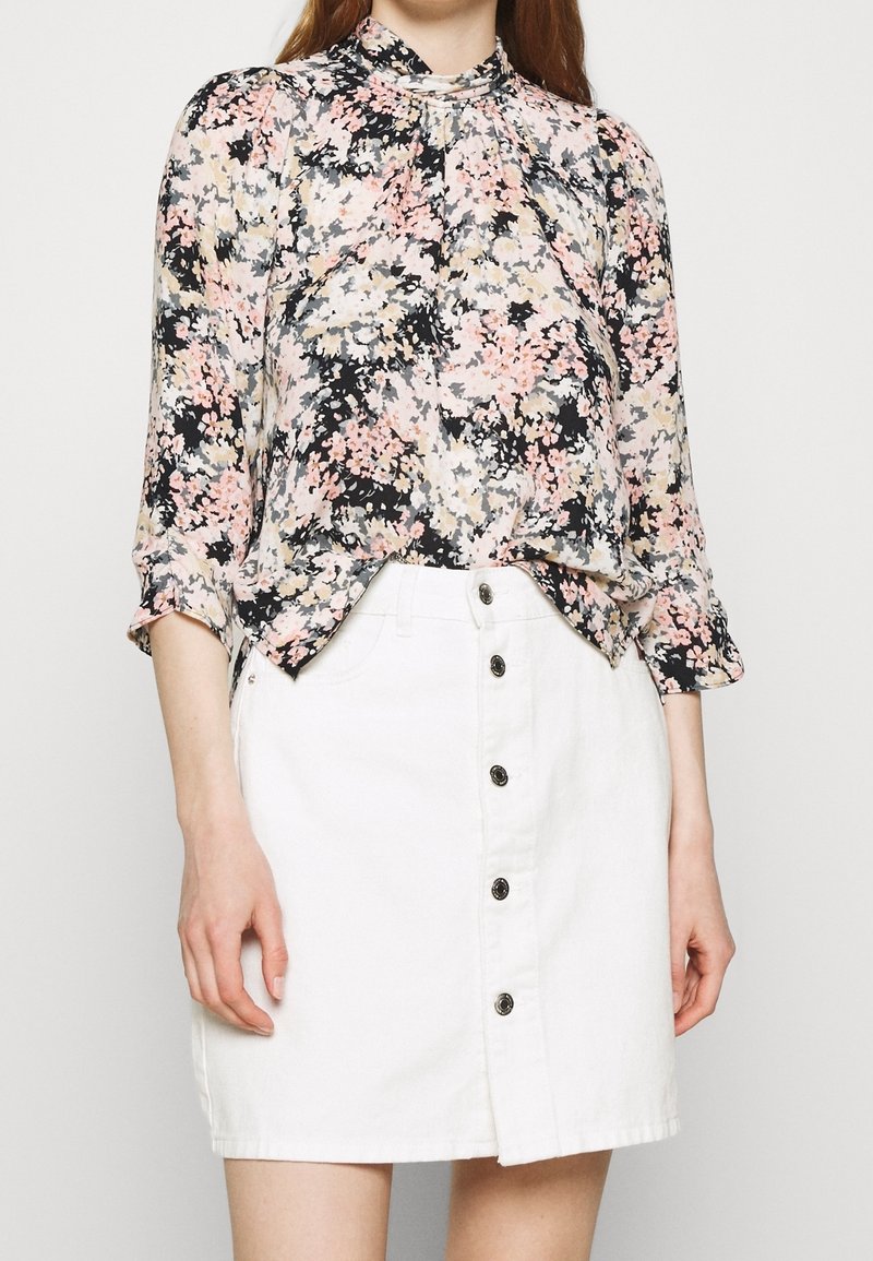 Blusa floreale in tessuto nero, rosa e crema con collo alto e maniche lunghe. Abbinata a una gonna in denim bianca con chiusura a bottoni.