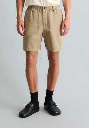 Mann mit beigefarbenen Shorts, schwarzen Socken und schwarzen Slippern, der vor einem schlichten hellen Hintergrund steht.