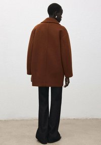 Bruine wollen jas met brede rever, oversized pasvorm en uitgesproken naden, gecombineerd met wijduitlopende zwarte broek. Gladde textuur en minimalistisch ontwerp.