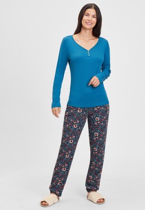 Blaue Langarm-Bluse mit V-Ausschnitt und Knopfakzenten, kombiniert mit gemusterten, floral bedruckten Schlafanzughosen in dunklen Tönen. Weicher, lässiger Schnitt.