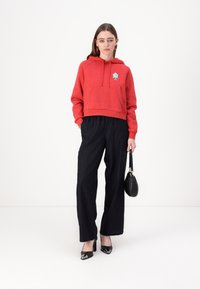 Red cropped hoodie with pocket and blue graphic, paired with black pinstripe wide-leg pants. Model holds a small black handbag.