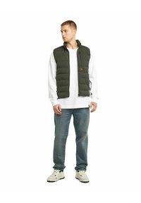 Olivgrön pufferväst med ståkrage, dragkedjestängning och kviltad design, lagd över en vit långärmad tröja och tillsammans med blå jeans.