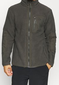 Mężczyzna w ciemnoszarej polarowej kurtce z zamkiem, zamek błyskawiczny na klatce piersiowej i wzmocnione opakowania na łokciach i ramionach.