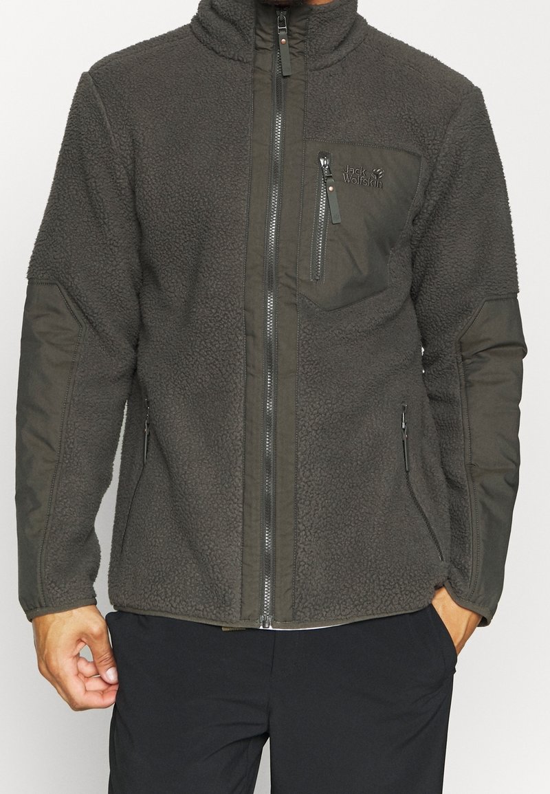 Mężczyzna w ciemnoszarej polarowej kurtce z zamkiem, zamek błyskawiczny na klatce piersiowej i wzmocnione opakowania na łokciach i ramionach.