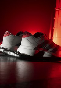 Weiße und rote Adidas Adizero Laufschuhe, die auf einer dunklen Oberfläche mit rot beleuchtetem Hintergrund ruhen.