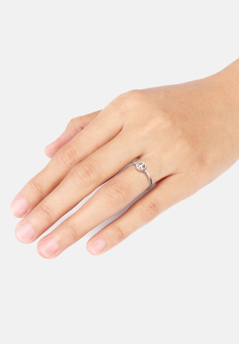 Silberner Frieden Zeichen Ring an einer Hand, mit einem dünnen Band und einem kreisförmigen Mittelpunkt mit einem Friedenssymbol-Design. Glatte Textur, minimalistischer Stil.