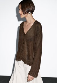 Bruine gebreide cardigan met een V-hals, lange mouwen en knoopsluiting. Gestructureerde stof met een losse pasvorm. Draagt dit over een lichte rok.