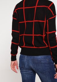 Person, der einen schwarzen Pullover mit roten Karos und blaue Jeans trägt, von hinten gezeigt, mit einer entspannten Hand an der Seite.