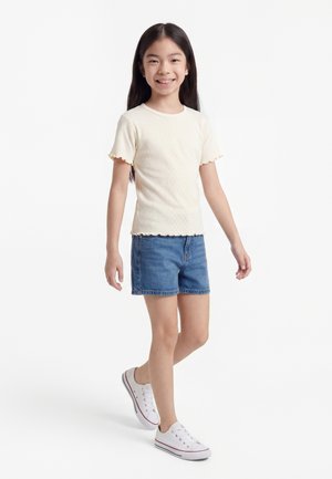 Ragazza sorridente che cammina, indossando una maglietta crema a maniche corte, pantaloncini di denim e sneakers bianche su uno sfondo bianco.