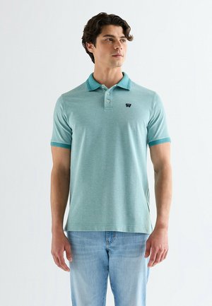 Mężczyzna w jasnooliwkowej, krótkim rękawie polo z kołnierzykiem oraz jasnoniebieskich dżinsach, stojący na tle jednolitego białego tła.