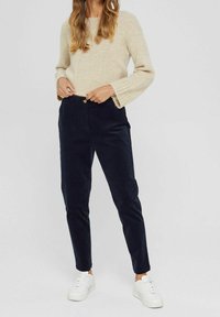 Beiger gestrickter Pullover mit weiten Ärmeln, kombiniert mit dunkelblauen Cordhosen im Slim-Fit-Stil mit Knopfverschluss, getragen mit weißen Sneakers.