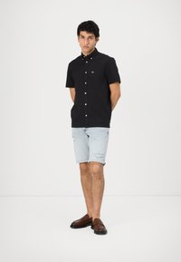 Camisa de botones negra de manga corta con un pequeño logo, combinada con pantalones cortos de denim desgastados en azul claro y mocasines marrones. Conjunto simple y casual.