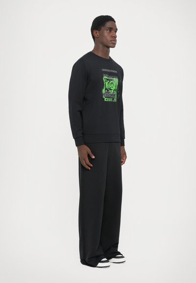 Schwarzes Sweatshirt mit langen Ärmeln und einem auffälligen grünen Grafikdesign. Kombiniert mit lockeren schwarzen Hosen und schwarz-weißen Turnschuhen.