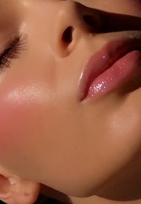 Close-up of a face featuring smooth skin, a subtle pink blush, glossy pink lips with sparkles, and a highlighted cheekbone.