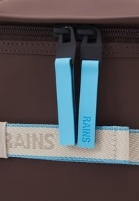 Fermeture éclair marron sur un sac avec deux tirettes en caoutchouc bleu étiquetées "RAINS" et une sangle en tissu beige avec bordure bleue.