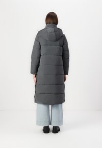 Grå polstret jakke med hætte, med vandrette quiltede sektioner, en mellemlang længde og split-sider, parret med lyseblå vide jeans.
