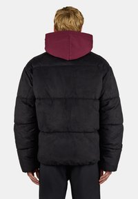 Blonde Person trägt eine schwarze gesteppte Jacke mit einer dunkelroten Kapuze und steht mit dem Rücken zu einem schlichten hellen Hintergrund.