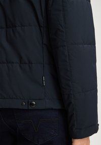 Granatowa kurtka puchowa z pikowanym wzorem, zapięcie na napy na dole, widoczna naszywka z logo z boku, noszona na ciemnych dżinsach z detalami w szwie.