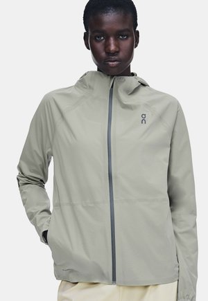 Personne portant une veste à capuche gris clair avec une fermeture éclair à l'avant et un logo sur la poitrine, debout avec une main dans une poche.