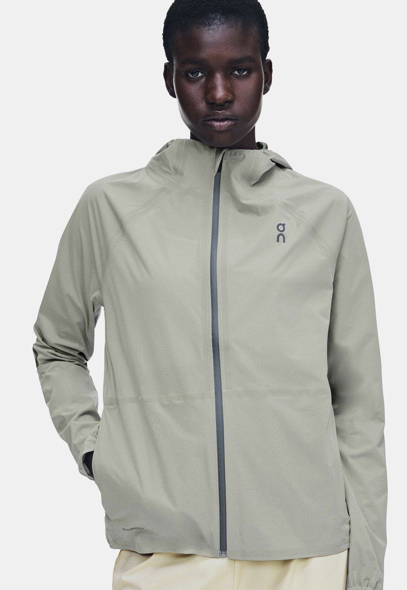 Personne portant une veste à capuche gris clair avec une fermeture éclair à l'avant et un logo sur la poitrine, debout avec une main dans une poche.