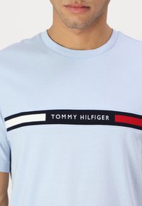 Camiseta de algodón azul claro con cuello redondo y una banda horizontal en azul marino que lleva el texto "TOMMY HILFIGER", acentuada por rayas rojas y blancas.