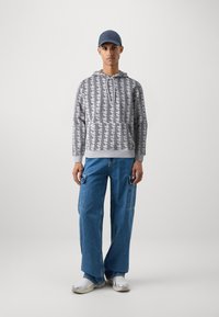 Felpa con cappuccio grigia chiara con un motivo geometrico nero, abbinata a pantaloni in denim blu larghi con tasche laterali. Sneakers bianche e un cappellino blu navy.