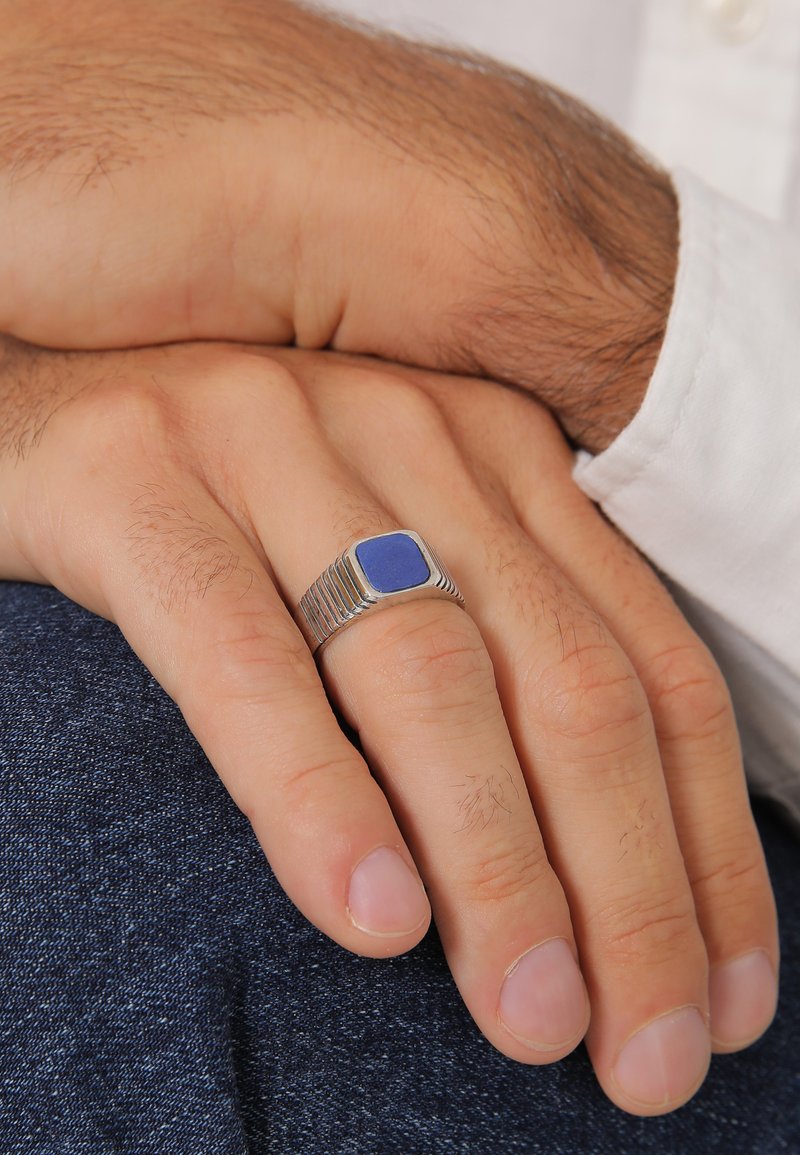 Anello d'argento con una pietra blu quadrata. Presenta dettagli a coste sulla banda. La mano è appoggiata su un tessuto in denim.
