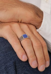 Sudraba gredzens ar kvadrātveida zilu akmeni. Joslai ir vertikālas rievas un pulēts apšuvums. Roka atpūšas uz džinsa auduma.