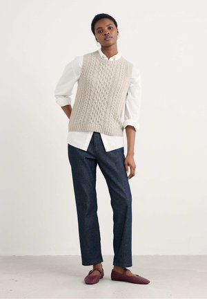 Licht beige gebreide vest met kabelpatronen, gelaagd over een wit shirt. Donkerdenim broek en kastanjebruine perforated flats maken de outfit compleet.