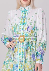 Robe à imprimé floral en matière blanche transparente, avec des manches bouffantes, des accents turquoise et une boucle de ceinture circulaire dorée. Boutons de couleur assortie.