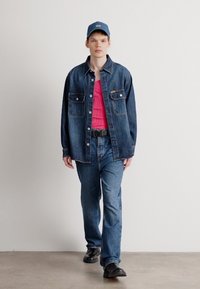 Τζάκετ από τζιν με κουμπωτή μπροστινή όψη, δύο τσέπες στο στήθος, ροζ πουκάμισο από κάτω και σκούρο τζιν. Μπλε καπέλο και μαύρα παπούτσια ολοκληρώνουν την εμφάνιση.