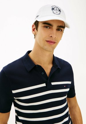 Jonge man met een witte pet met logo en een donkerblauw poloshirt met witte horizontale strepen, poseert tegen een effen witte achtergrond.