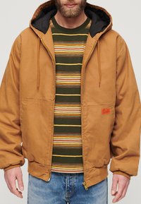 Chaqueta capucha marrón clara con cremallera frontal, parche en el pecho y dos bolsillos laterales. Llevada sobre una camiseta a rayas verde y marrón clara. Jeans azules visibles en la parte inferior.