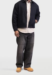 Czarna kurtka z zamkiem z przodu i kieszenią na piersi na jasnej koszuli zapinanej na guziki; do tego ciemnoszare, luźne dżinsy i beżowe drewniaki.