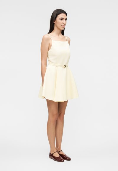 Γυναίκα που φοράει μια κρεμ αμάνικη φόρεμα με ζώνη, συνδυασμένη με σκούρα καφέ παπούτσια Mary Jane, σε ένα απλό φωτεινό φόντο.