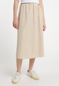 Beige midi szoknya vertikális piros csíkokkal, rugalmas derékpánttal, elülső gombos záródással és oldalsó zsebekkel, fehér sportcipővel párosítva.
