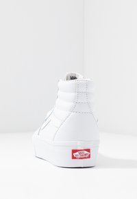 Vit hög sneakers i läder med vadderad ankel, med en grå sidrand och en röd Vans "Off the Wall"-logotyp på sulan.