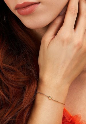 Close-up of a hand resting near the face, showing a delicate gold bracelet with a small heart charm on the wrist.