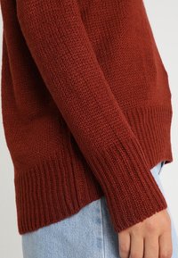 Dzianinowy sweter w kolorze rdzy z prążkowanymi mankietami i ściągaczem. Charakteryzuje się fakturowanym wzorem i luźnym krojem, noszony na jasnych dżinsach.