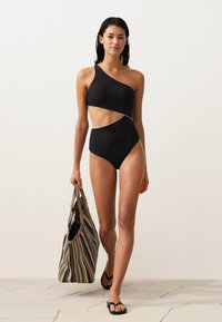 Femeie care merge purtând un costum de baie negru cu un umăr gol și decupaje, ținând o geantă mare cu dungi și purtând șlapi negri pe un podea deschisă la culoare.