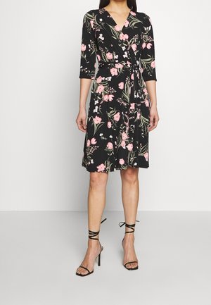 Femme portant une robe portefeuille noire à imprimé floral rose, manches trois-quarts, et des sandales à talons hauts noires avec lanières, sur un fond uni.