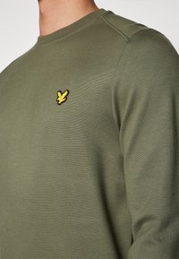 Suéter verde oliva hecho de algodón suave, con cuello redondo y un pequeño logotipo amarillo bordado en el pecho. Textura suave.