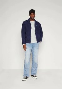 Ung man som bär en marinblå jacka, ljusgrå t-shirt, ljusblå jeans, svart keps och svart-vita sneakers, står framför en enkel bakgrund.