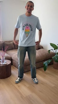 Νέος άνδρας χαμογελά και προσαρμόζει ένα ανοιχτό μπλε μπλουζάκι με πολύχρωμο παγωτό σε χωνάκι και το κείμενο «Always Fresh», όρθιος σε ένα σαλόνι.