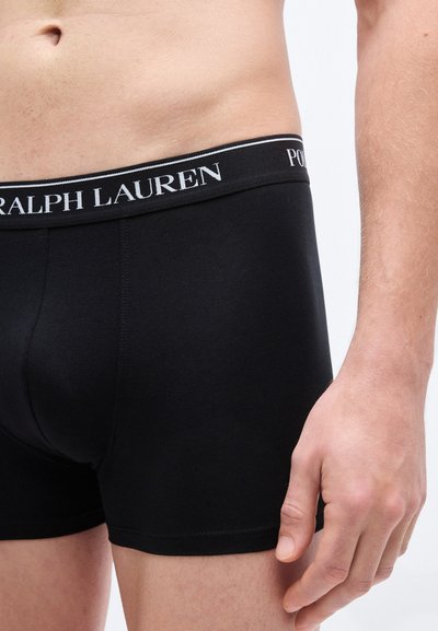 Crne bokserice s širokim pojasom na kojem je bijeli logo "Ralph Lauren". Tkanina izgleda glatko i prijanja uz tijelo.