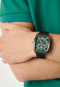 Reloj de pulsera negro con caja rectangular, acentos verdes, dial transparente y correa de goma, mostrado en una mano contra una camisa verde.