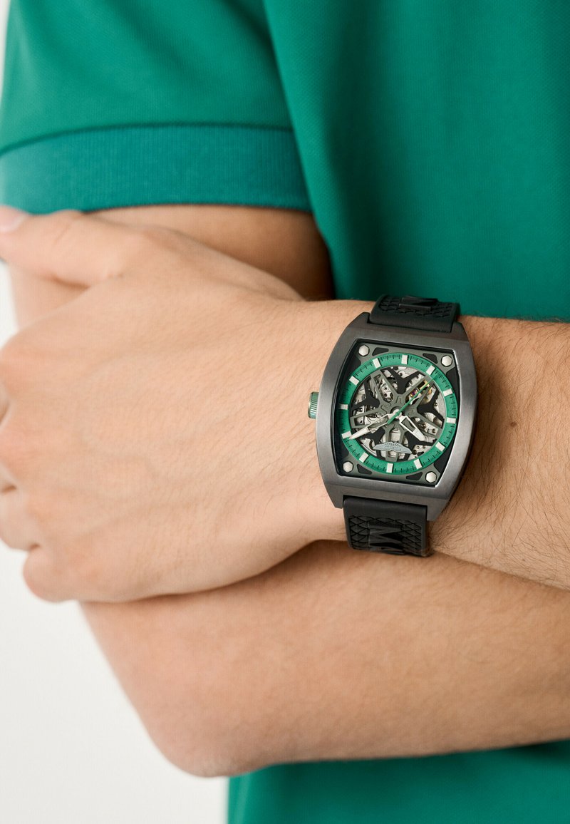 Reloj de pulsera negro con caja rectangular, acentos verdes, dial transparente y correa de goma, mostrado en una mano contra una camisa verde.