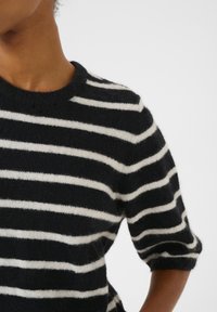 Černobílý pruhovaný svetr, vyrobený z měkkého materiálu, má kulatý výstřih a středně dlouhé rukávy s mírně chlupatou texturou.