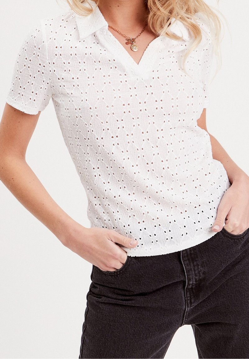 Femme portant une blouse blanche à manches courtes en broderie anglaise avec col et un pantalon noir, mains posées négligemment dans les poches avant.
